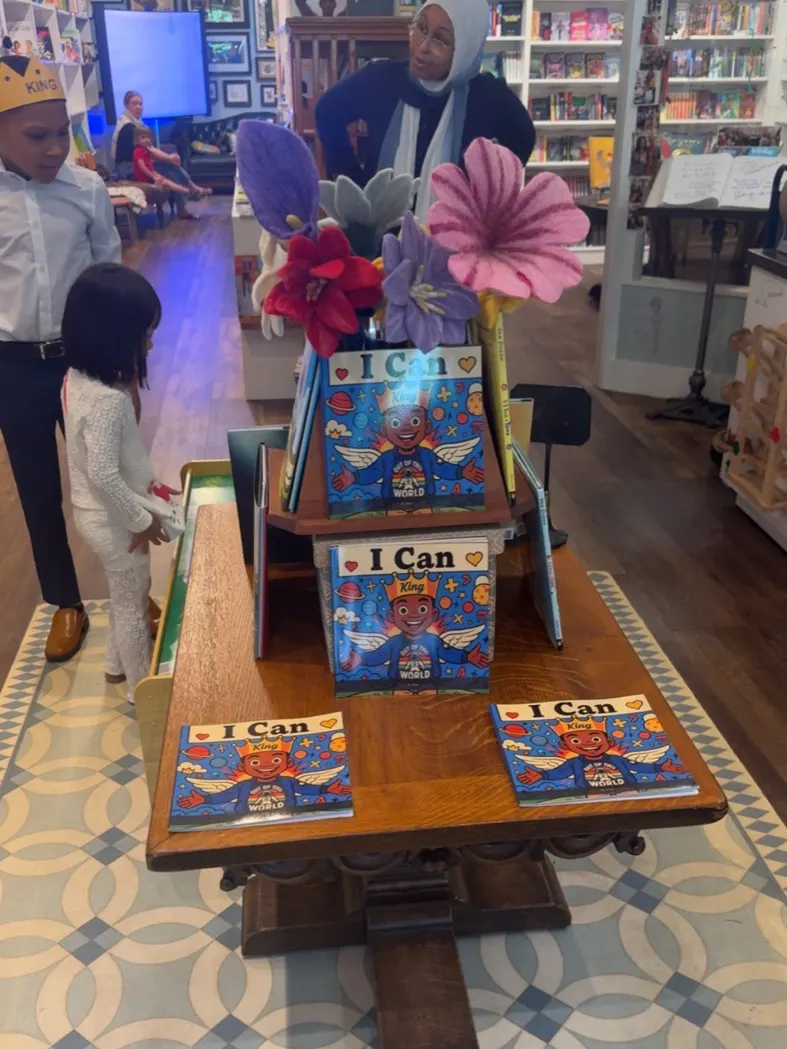Copies of I Can displayed on a wooden table in the bookstore, with felt flowers behind them and Titan standing at the edge of the frame in his crown.