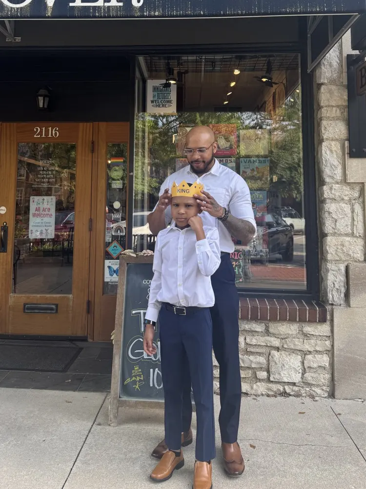 Titan's dad setting the gold paper KING crown on his head outside the Cover to Cover storefront, the event chalkboard beside them.